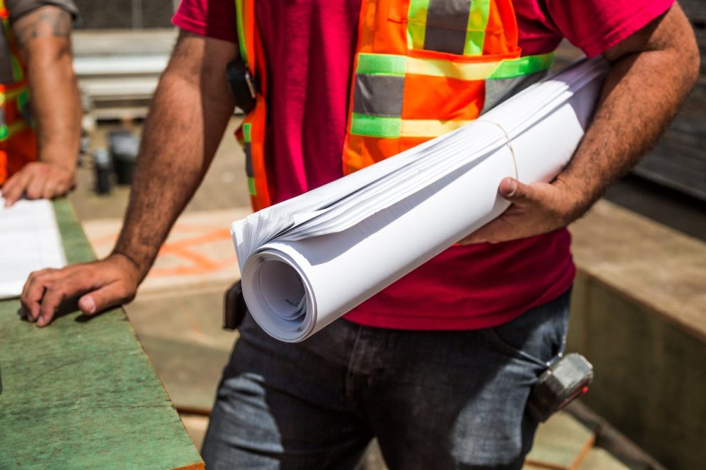 A male construction worker wearing an orange vest and red shirt holding a roll of construction plans on his left hand and his right hand is resting on the green table in front of him. A measuring tape is on the left side pocket of the construction worker.