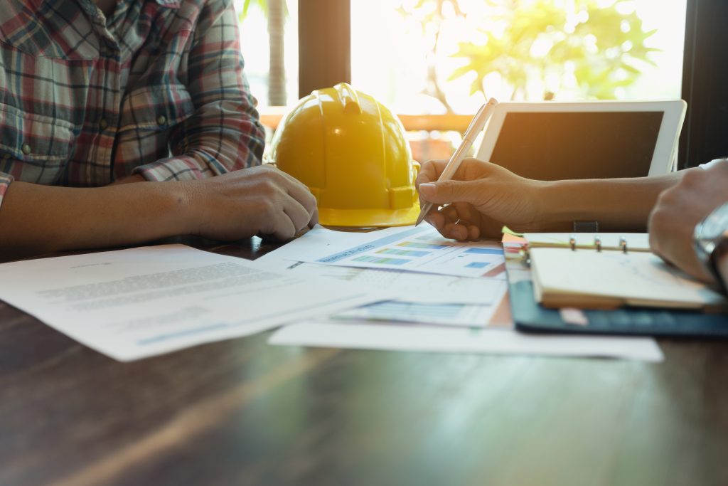 A contractor and customer discussing business plans. A yellow hard hat and tablet is on top of the wooden table.