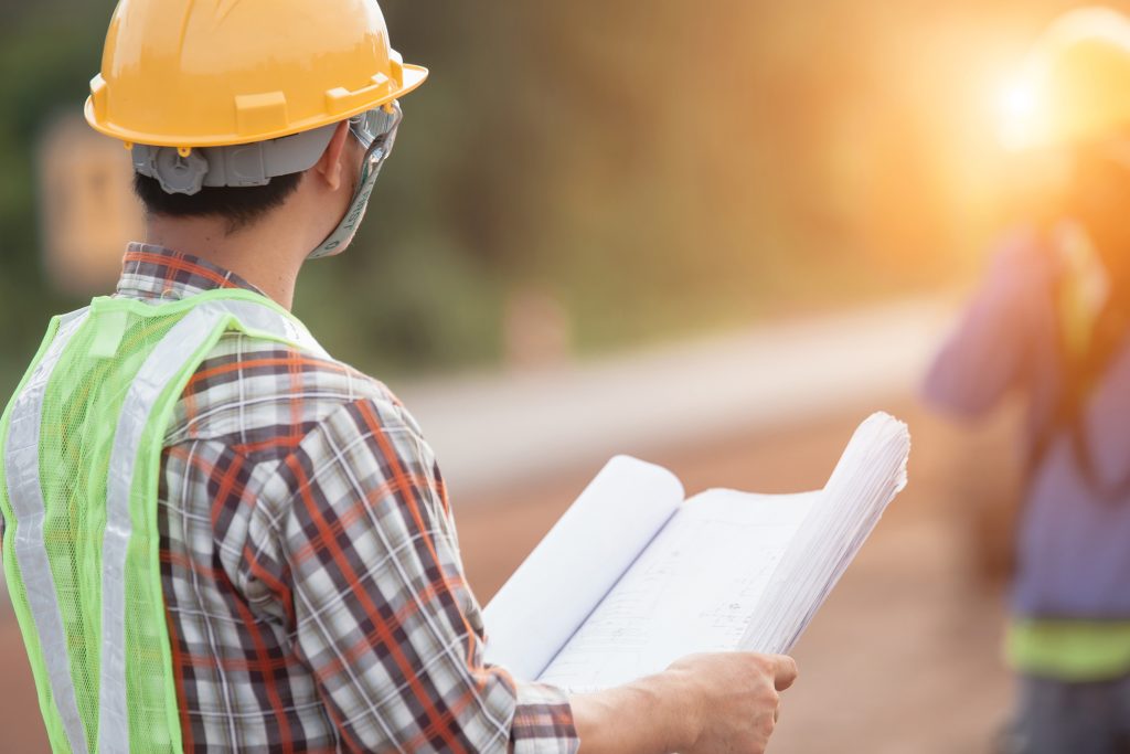 A construction worker wearing a yellow hard hat and green vest is holding a blueprint. A silhouette of another construction worker can be seen against the sunlight.