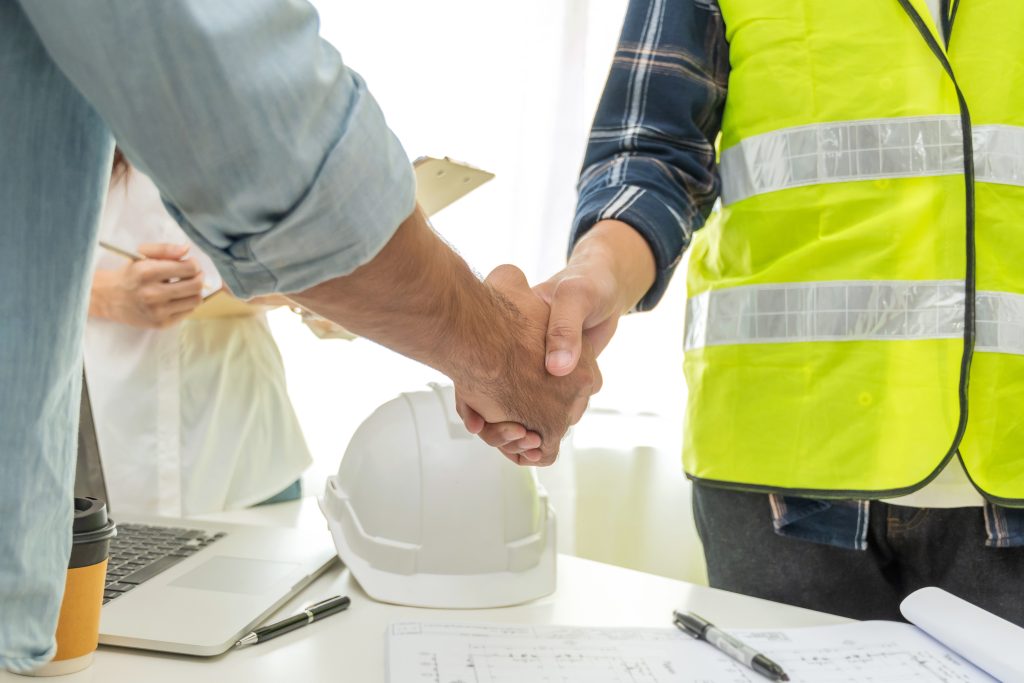 A contractor wearing green vest handshakes with a customer after business meeting. A laptop, coffee cup, pen, hard hat, and a blueprint is on the desk. A secretary holding a pen and folder wearing a white shirt is on the left side.