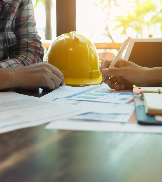 A contractor and customer discussing business plans. A yellow hard hat and tablet is on top of the wooden table.