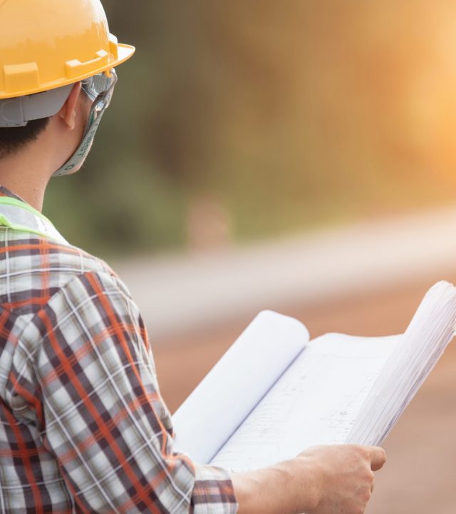 A construction worker wearing a yellow hard hat and green vest is holding a blueprint. A silhouette of another construction worker can be seen against the sunlight.